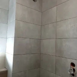 Ściana w łazience wyłożona szarymi, matowymi płytkami ceramicznymi, widoczne otwory na wentylację i gniazdka elektryczne w trakcie montażu.