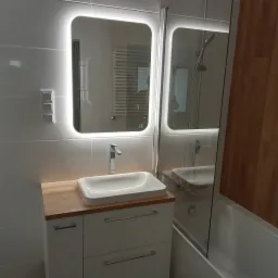 Nowoczesna łazienka z białymi płytkami, umywalką nablatową na szafce z drewnianym blatem i podświetlanym lustrem z zaokrąglonymi rogami, widoczna kabina prysznicowa.