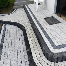 Ułożona kostka brukowa w kolorach szarości prowadząca do wejścia domu, z obrzeżami z ciemnych, okrągłych elementów.