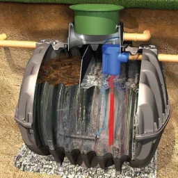 Przekrój szamba ekologicznego z widocznymi komorami, filtrami i instalacją pompy, umieszczonego w ziemi na podsypce z kruszywa.