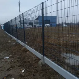 Nowo postawione, panelowe ogrodzenie z podmurówką betonową na grząskim terenie, za nim budynek przemysłowy i zaparkowane busy, pochmurne niebo.