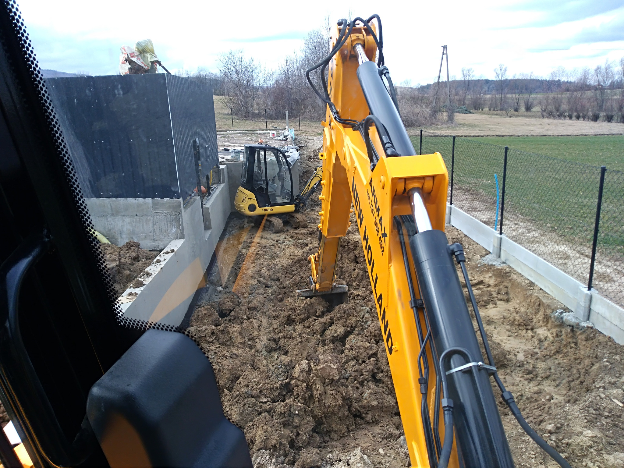 Żółta koparka New Holland podczas prac ziemnych pod fundamenty w Łużnej, widok z kabiny drugiej maszyny.