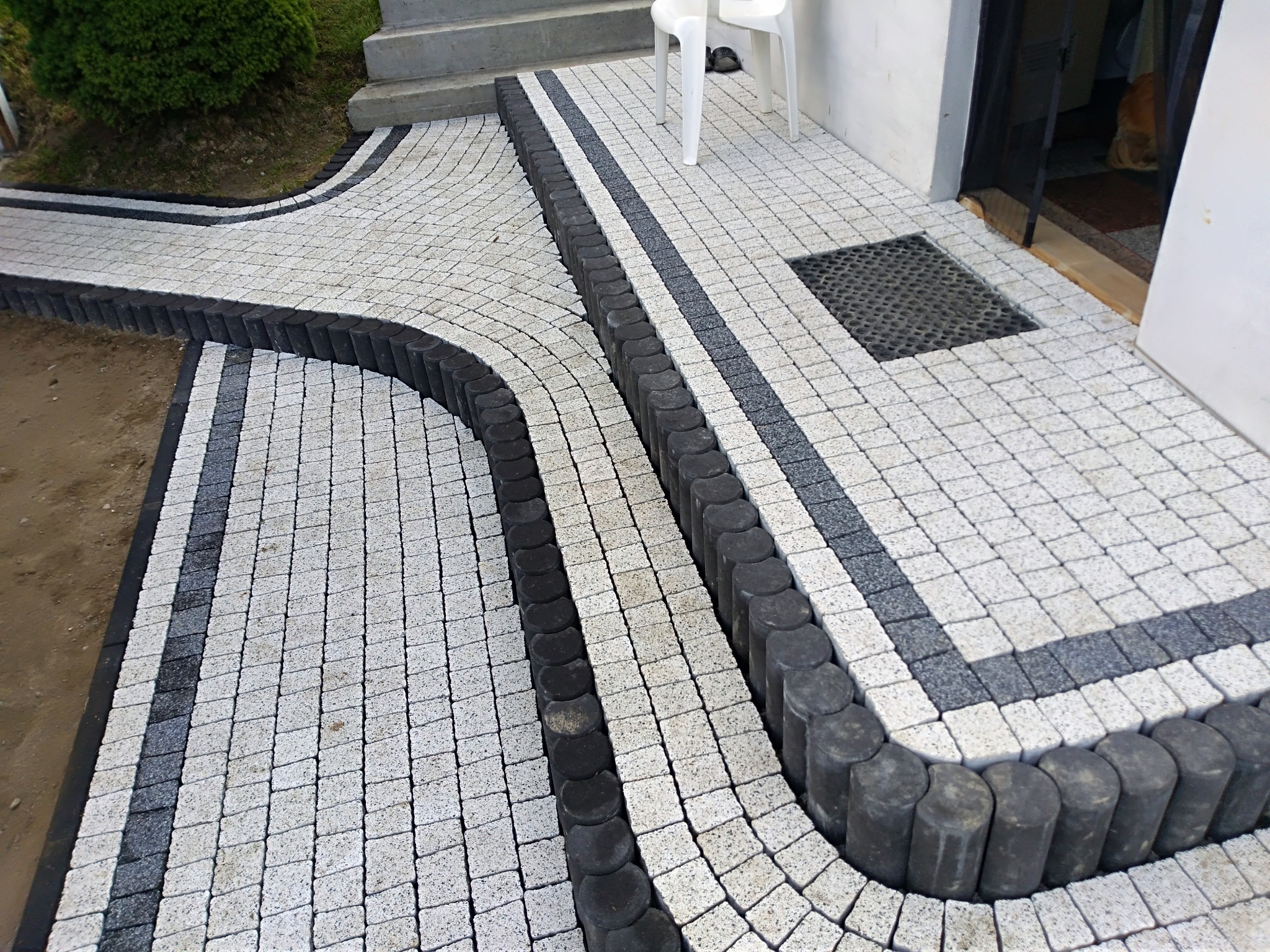 Ułożona kostka brukowa w kolorach szarości prowadząca do wejścia domu, z obrzeżami z ciemnych, okrągłych elementów.
