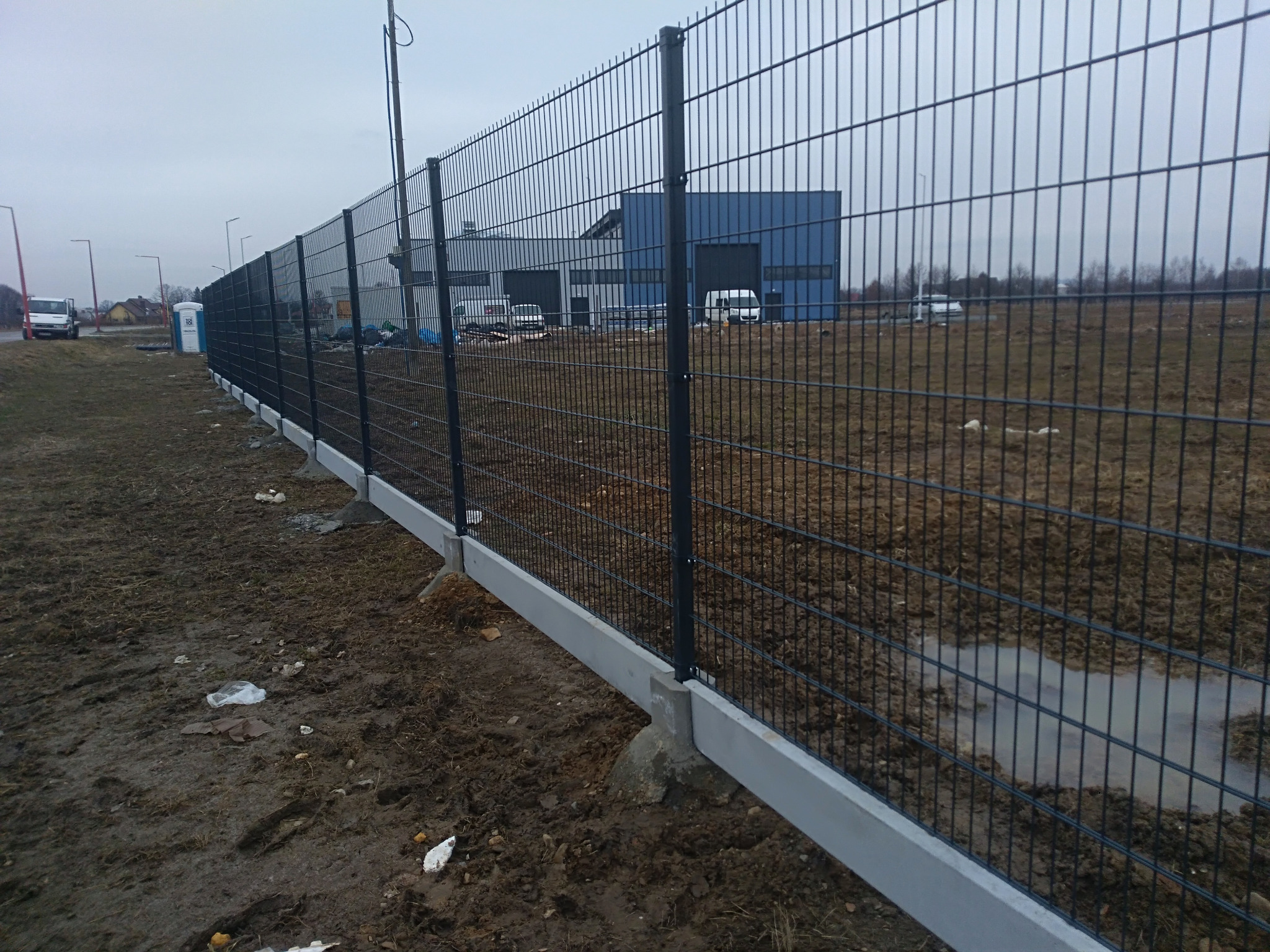 Nowo postawione, panelowe ogrodzenie z podmurówką betonową na grząskim terenie, za nim budynek przemysłowy i zaparkowane busy, pochmurne niebo.