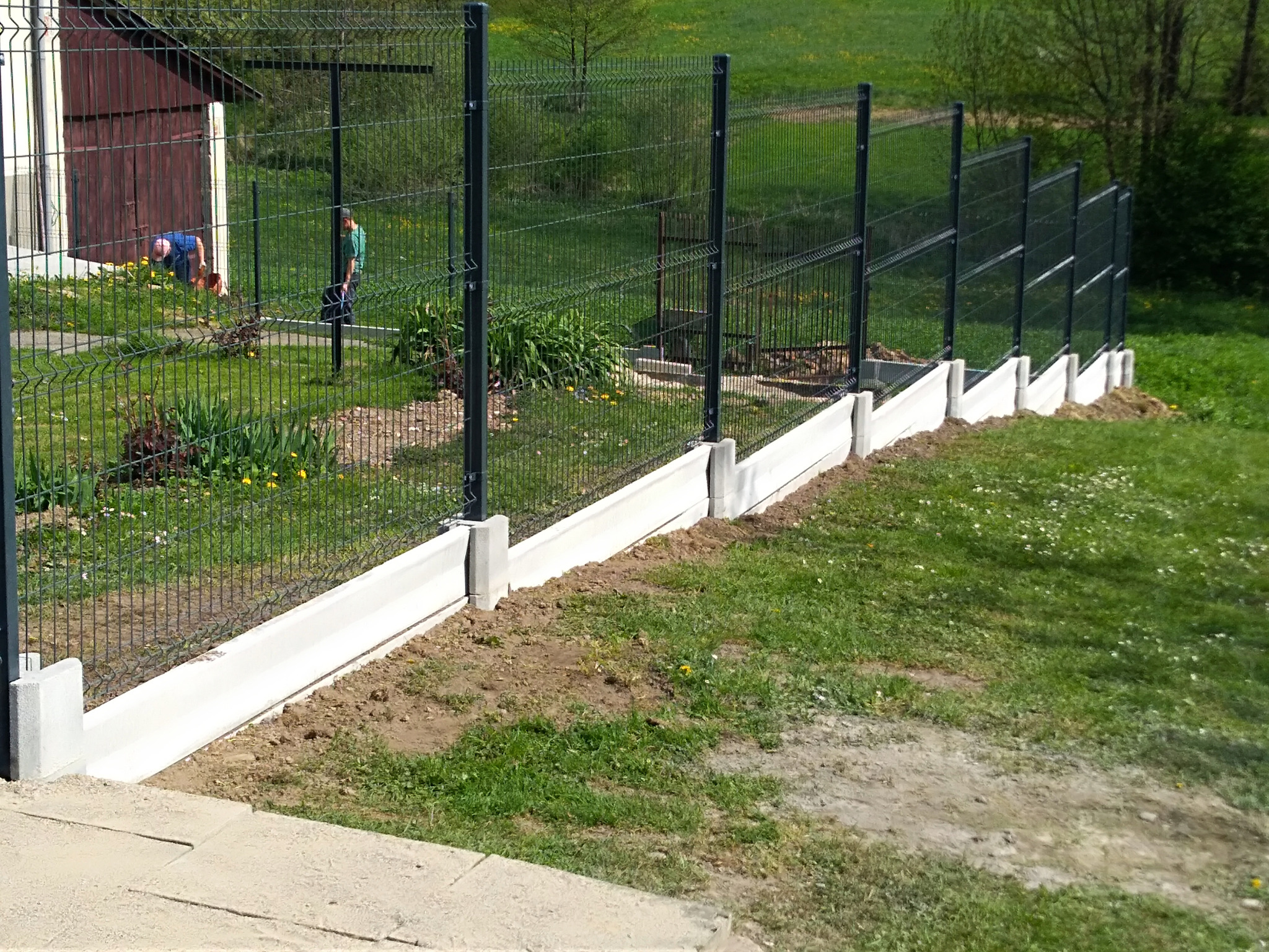 Nowoczesne ogrodzenie panelowe z podmurówką betonową na trawiastym terenie; w tle budynek gospodarczy i osoby pracujące w ogrodzie.
