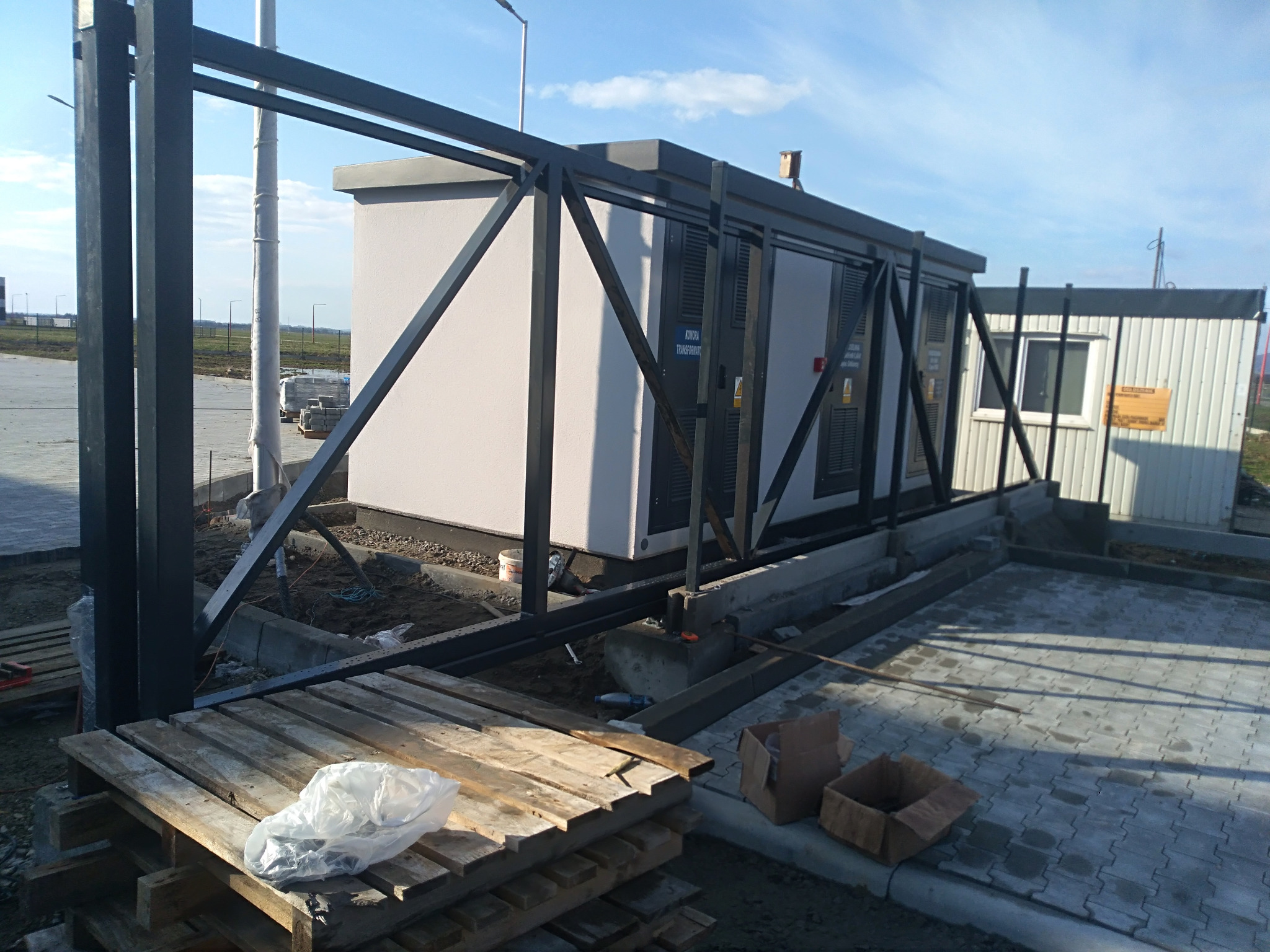 Montaż prefabrykowanej komory transformatorowej na betonowym fundamencie, widoczny metalowy szkielet konstrukcyjny, palety i kostka brukowa w tle, teren budowy.