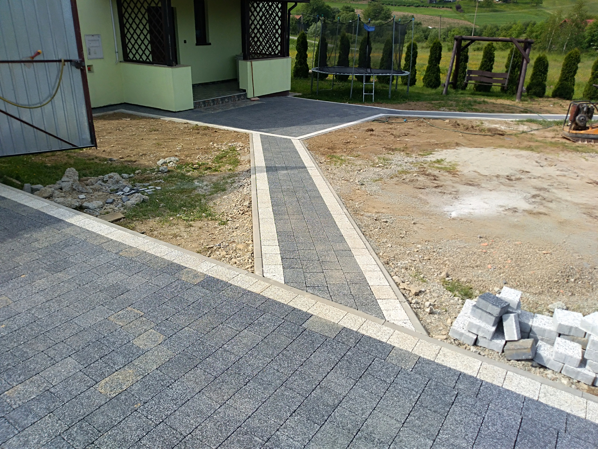 Świeżo ułożona, szara kostka brukowa z jasnymi obrzeżami tworząca ścieżkę i podjazd przed domem z werandą, w tle trampolina i huśtawka na tle zielonego wzgórza.