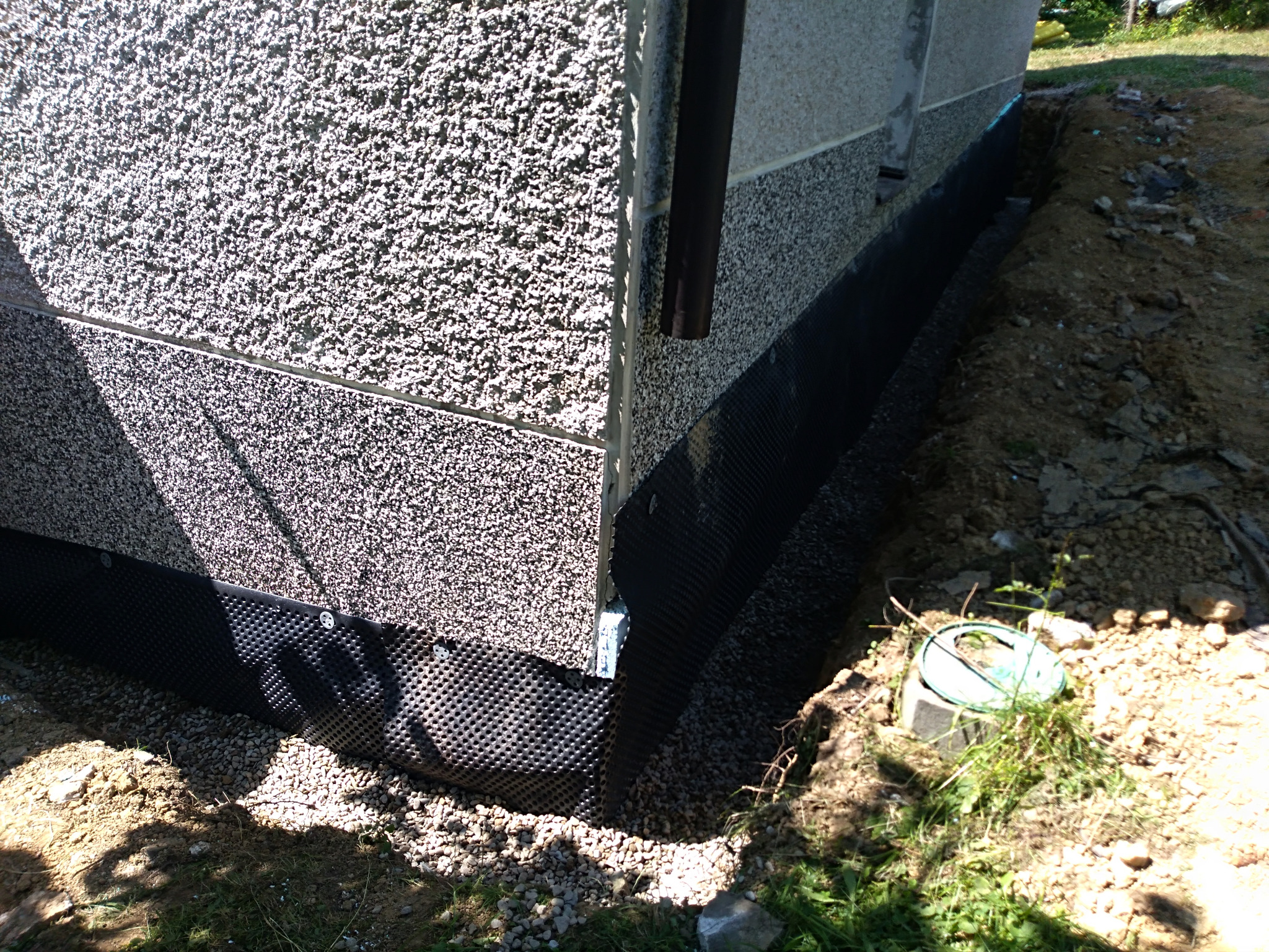 Izolacja fundamentu budynku z użyciem folii kubełkowej, widoczne mocowania, zasypany drenaż z rurą inspekcyjną. Ściana budynku z tynkiem strukturalnym.