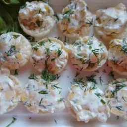 Kruche babeczki ryżowe wypełnione sałatką z majonezem, kukurydzą i posypane koperkiem, ułożone na białym talerzu z liśćmi sałaty.