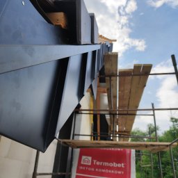 Dawid Śmiertka - Szczegółowe ujęcie czarnej, metalowej obróbki blacharskiej na krawędzi dachu, z widocznym rusztowaniem i fragmentem ściany budynku w tle.