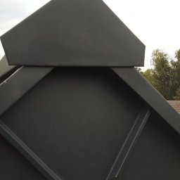 Dawid Śmiertka - Szczegółowe ujęcie szarego, metalowego wykończenia dachu w kształcie piramidy, z widocznymi liniami łączeń i fragmentem tła z zielenią drzew.