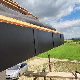 Dawid Śmiertka - Czarny panel okapowy zamontowany na dachu z widoczną membraną i elementami drewnianymi, na tle zielonego krajobrazu i błękitnego nieba z chmurami.