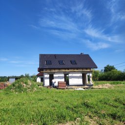 Dawid Śmiertka - Dom w trakcie budowy z czarnym dachem z dachówkami i oknami dachowymi, na tle błękitnego nieba z chmurami. Widoczny pracownik i materiały budowlane.