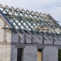 Dawid Śmiertka - Surowa więźba dachowa z impregnowanego drewna na budynku z białych bloczków, widok z dołu na tle jasnego nieba. Konstrukcja w trakcie montażu, widoczne elementy mocujące.