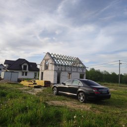 Dawid Śmiertka - Dom w trakcie budowy z widoczną więźbą dachową i zaparkowanym czarnym Mercedesem. W tle drugi dom i składowane drewno budowlane.