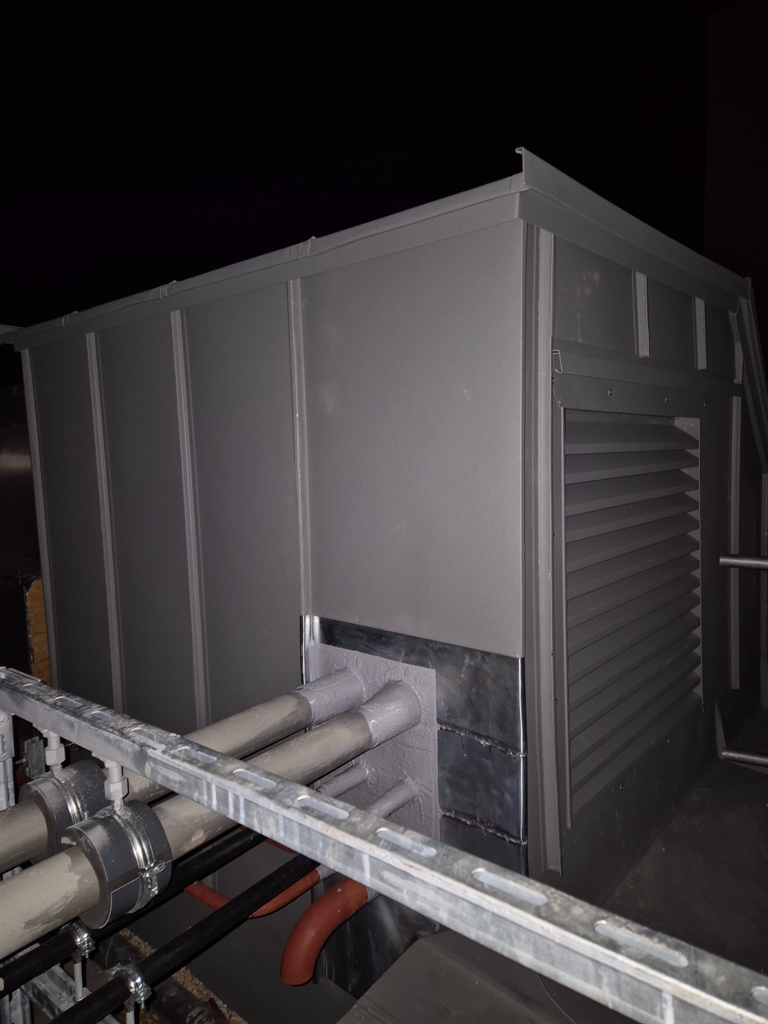 Szara, metalowa obudowa na dachu z widocznymi rurami i elementami wentylacyjnymi. Nocne ujęcie z bliska, ukazujące detale konstrukcji i połączenia.