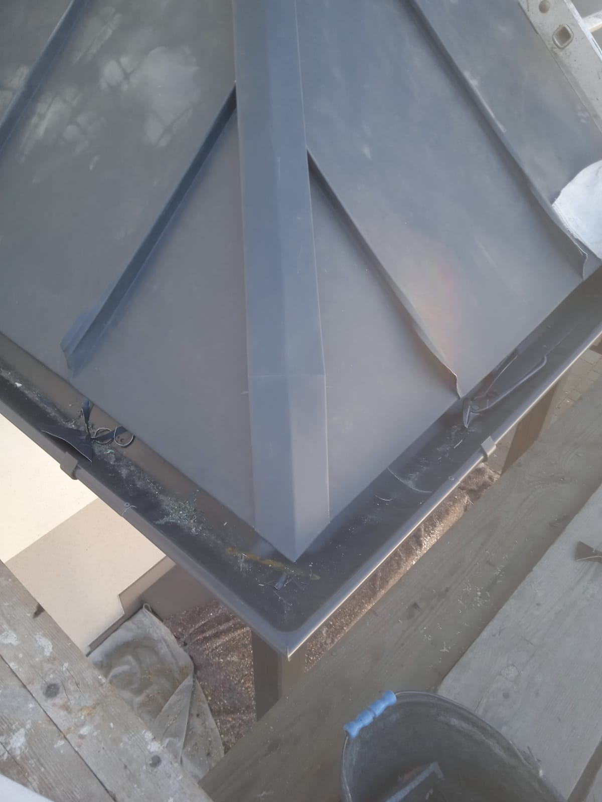 Szary panel dachowy z elementami mocującymi, widok z góry, z bliska. Fragment rynny z drobnymi zanieczyszczeniami. Widoczne drewniane elementy konstrukcyjne.