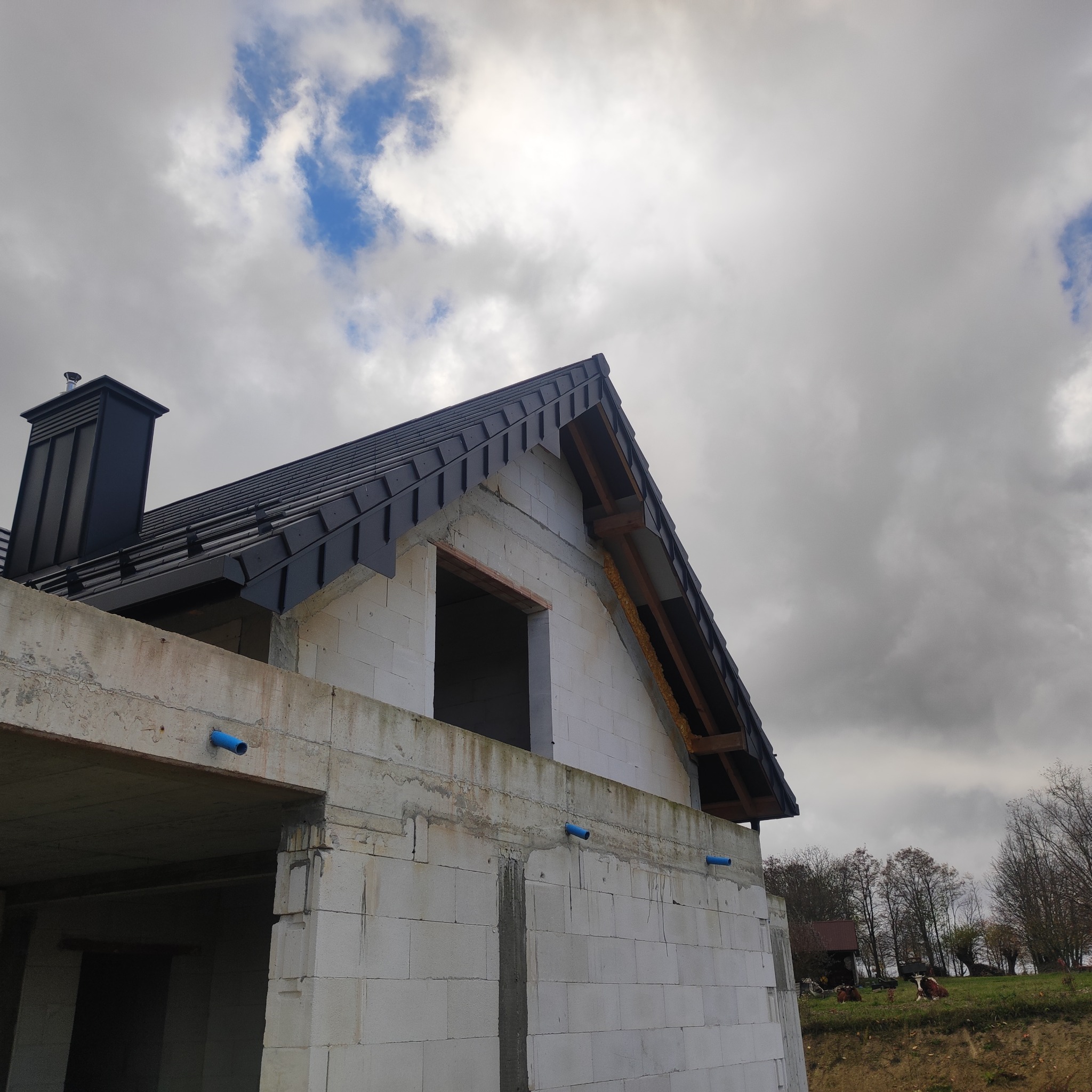 Dom w trakcie budowy z czarnym dachem i kominem. Widoczne niezadaszone okno, ściany z białych bloczków i fragment krajobrazu z pochmurnym niebem w tle.