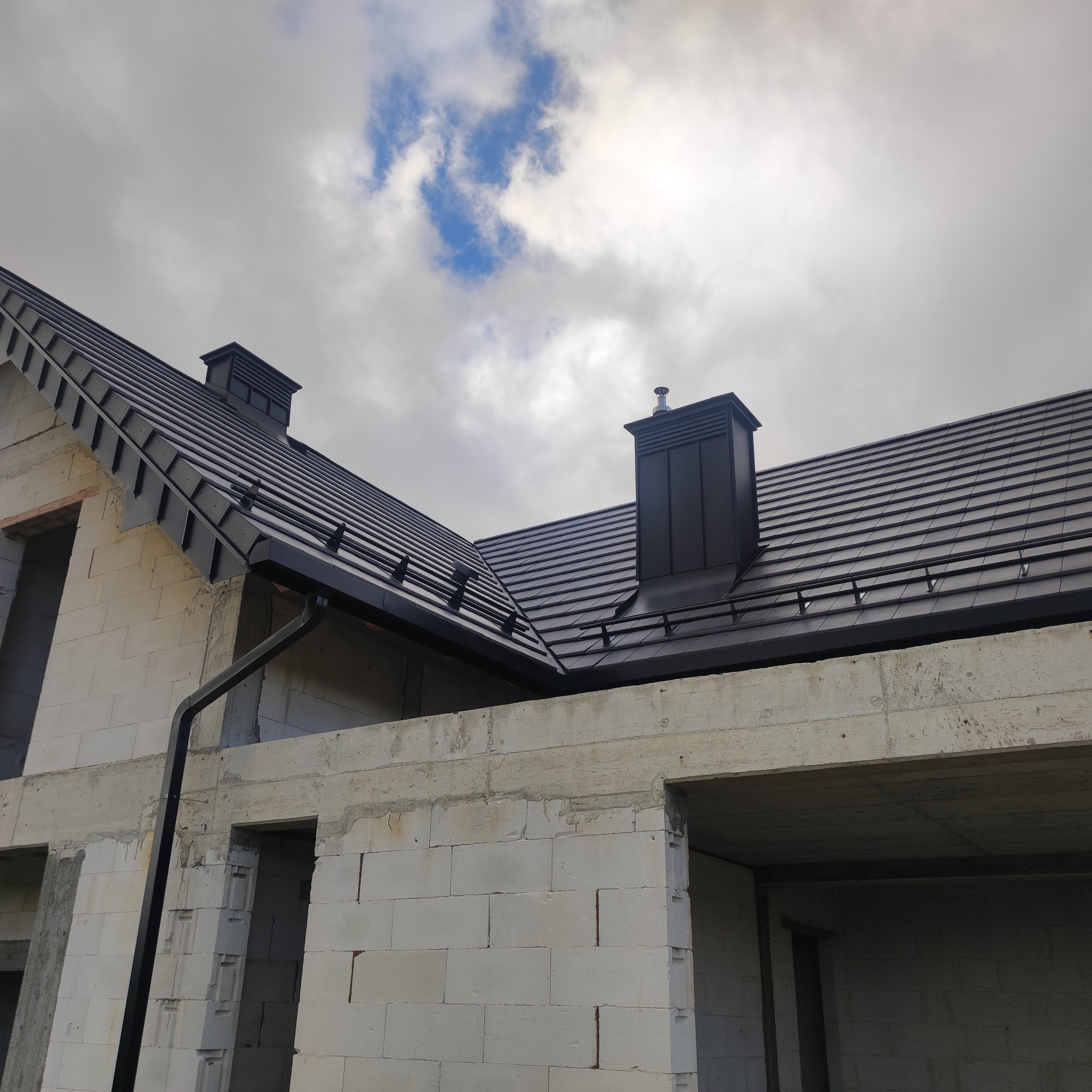 Nowoczesny dach z czarnej dachówki ceramicznej i obróbkami blacharskimi na surowym budynku z białego betonu komórkowego w pochmurny dzień.