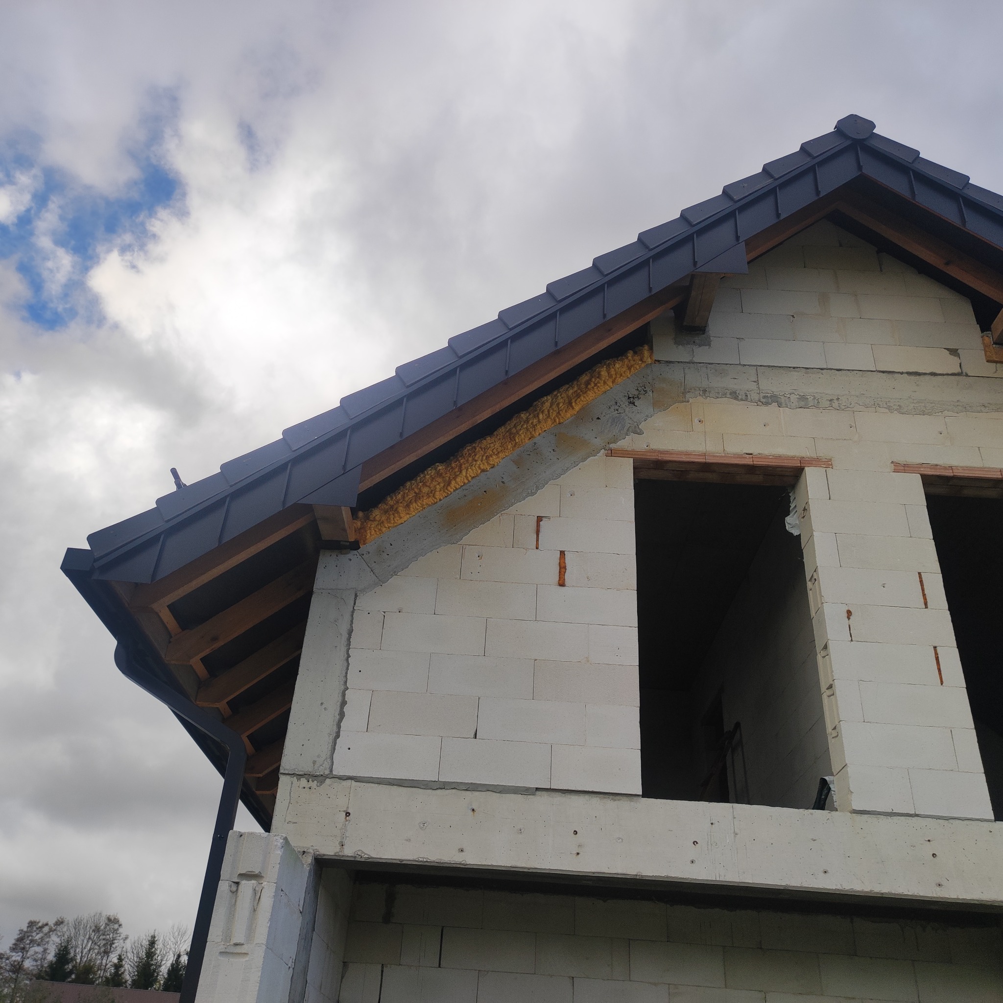 Surowy stan budynku z widoczną konstrukcją dachu z czarną dachówką i ociepleniem pianką. Widoczne niezadaszone okna i szare niebo w tle.