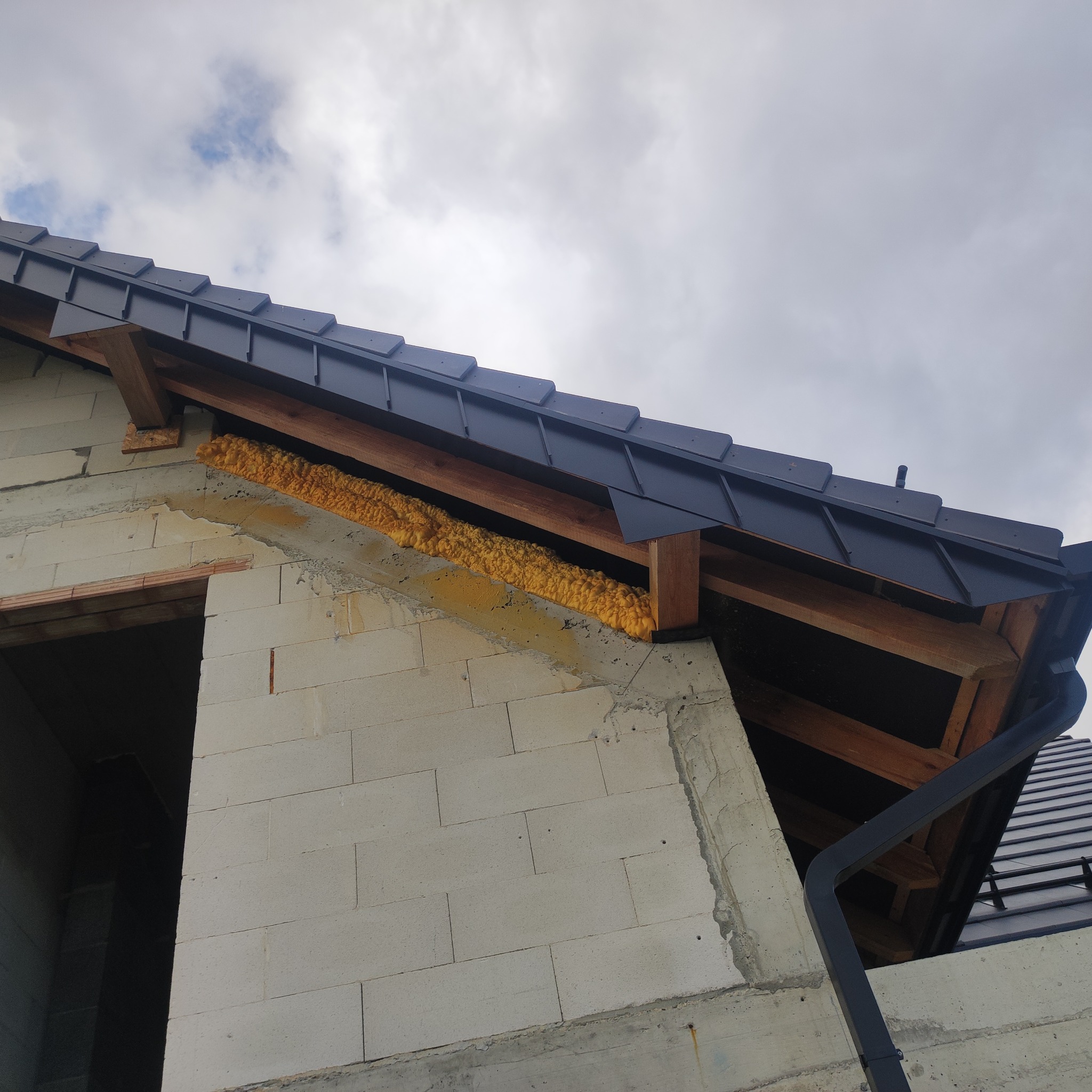 Szczegółowe ujęcie dachu z czarną blachodachówką i pianką izolacyjną w Tarnowie. Widoczna więźba dachowa i fragment rynny. Ujęcie z dołu, pochmurne niebo w tle.