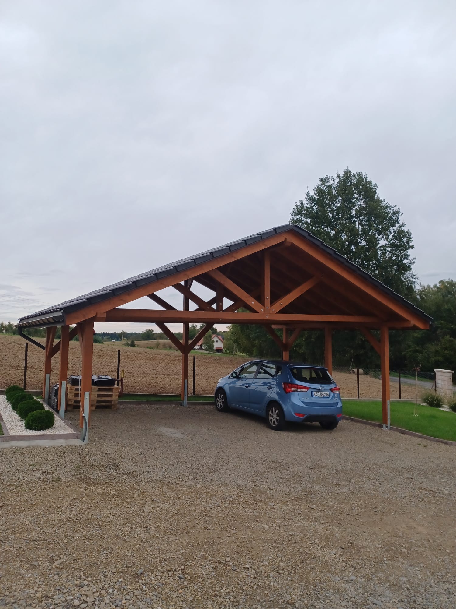 Drewniana wiata garażowa z ciemną dachówką, pod którą stoi niebieski samochód. W tle pole i drzewa, całość w pochmurny dzień.