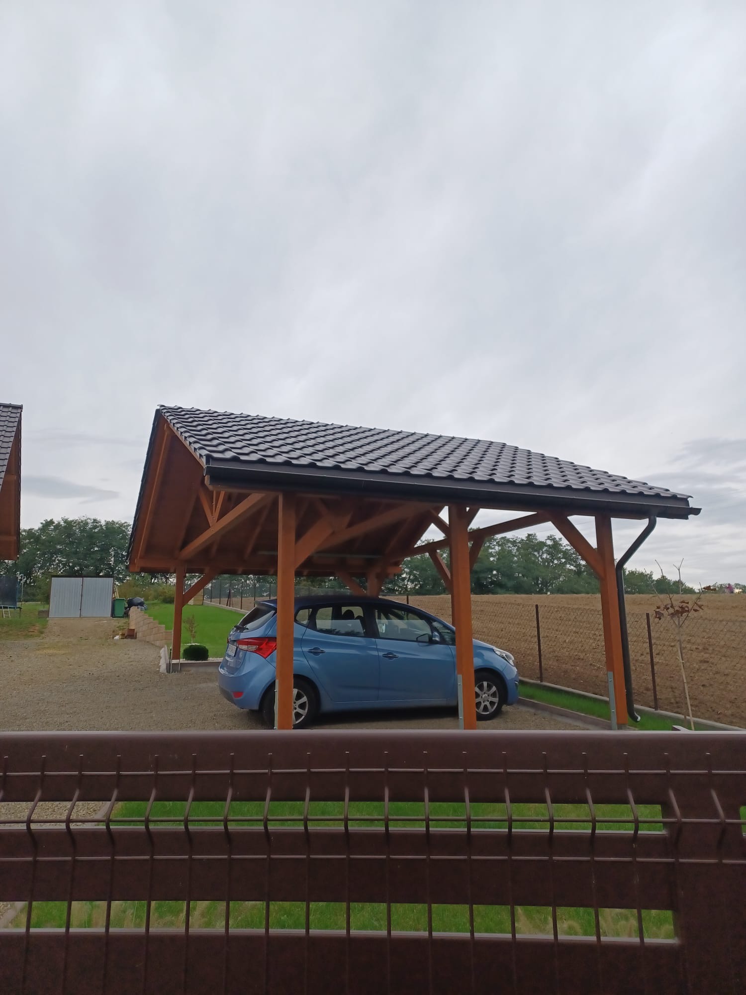 Drewniana wiata garażowa z ciemną dachówką kryjąca błękitny samochód, widok zza brązowego ogrodzenia. W tle zielona trawa i pochmurne niebo.