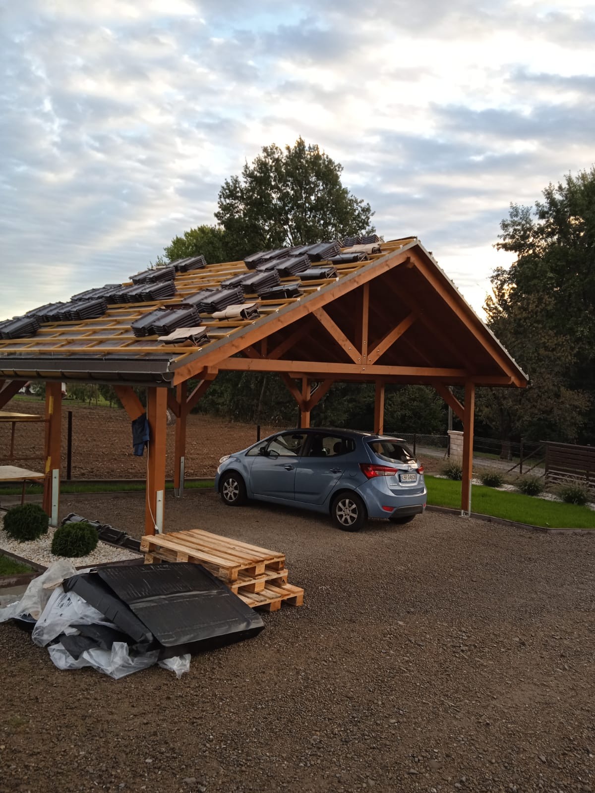 Drewniana wiata z częściowo ułożoną dachówką ceramiczną, obok zaparkowany niebieski samochód. Widoczne palety i materiały budowlane. Prace dekarskie w toku.