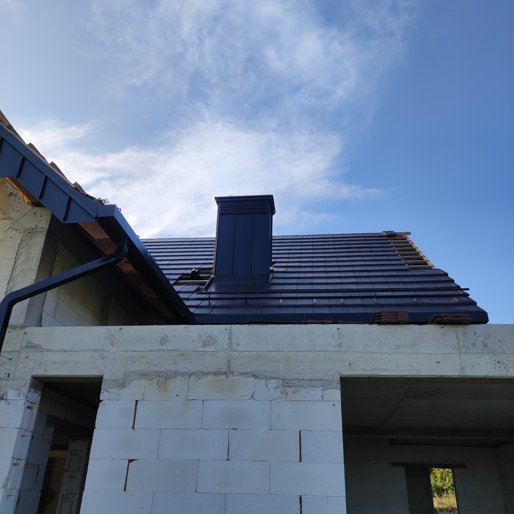 Niski kąt na dom w budowie z czarną dachówką i kominem. Widoczne rynny i fragment więźby. Błękitne niebo w tle.