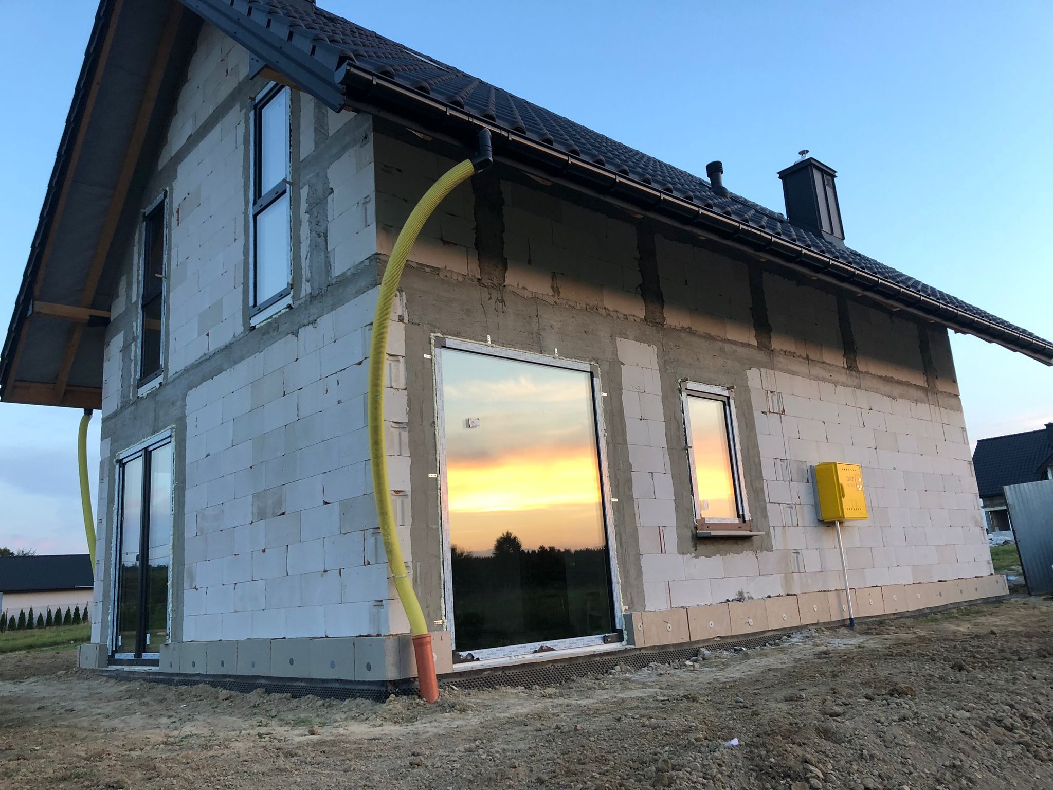 Surowy stan domu jednorodzinnego z czarnym dachem i rynnami, widoczne okna z odbiciem zachodu słońca, żółta rura i skrzynka gazowa na elewacji.