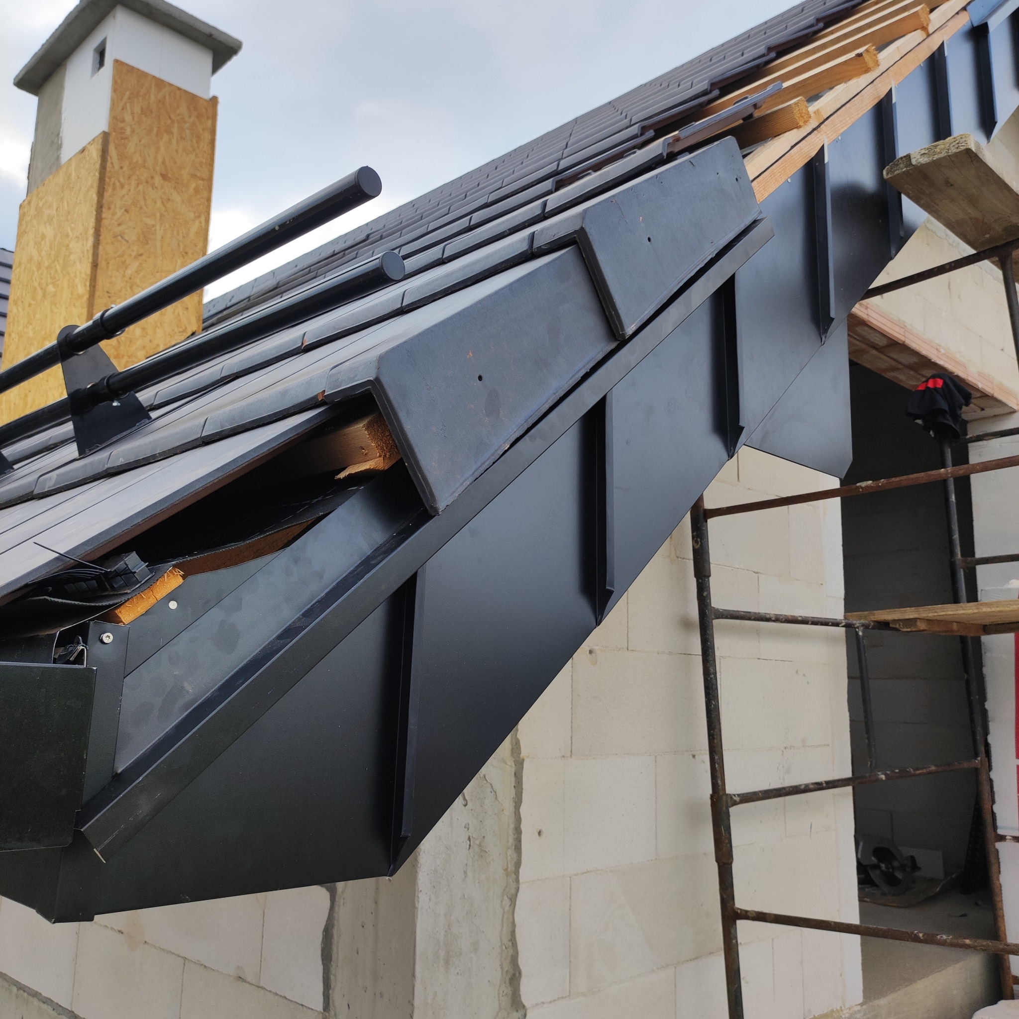 Czarny, metalowy system orynnowania na dachu z dachówką, widoczny z dołu pod kątem. Fragment rusztowania i komina w tle. Detal konstrukcji dachu.