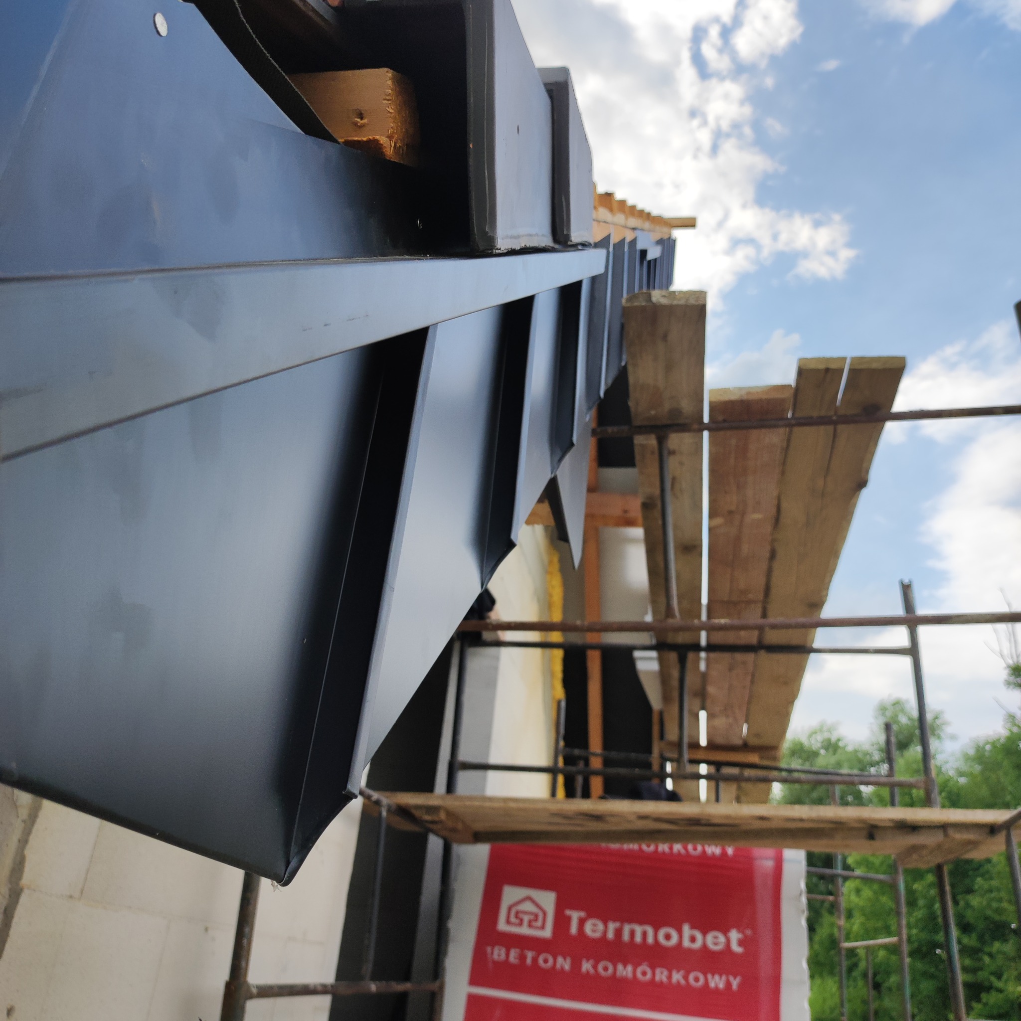 Szczegółowe ujęcie nowej, błyszczącej, czarnej obróbki blacharskiej dachu z blachy na tle rusztowania i błękitnego nieba. Widoczny fragment ściany z betonu komórkowego.