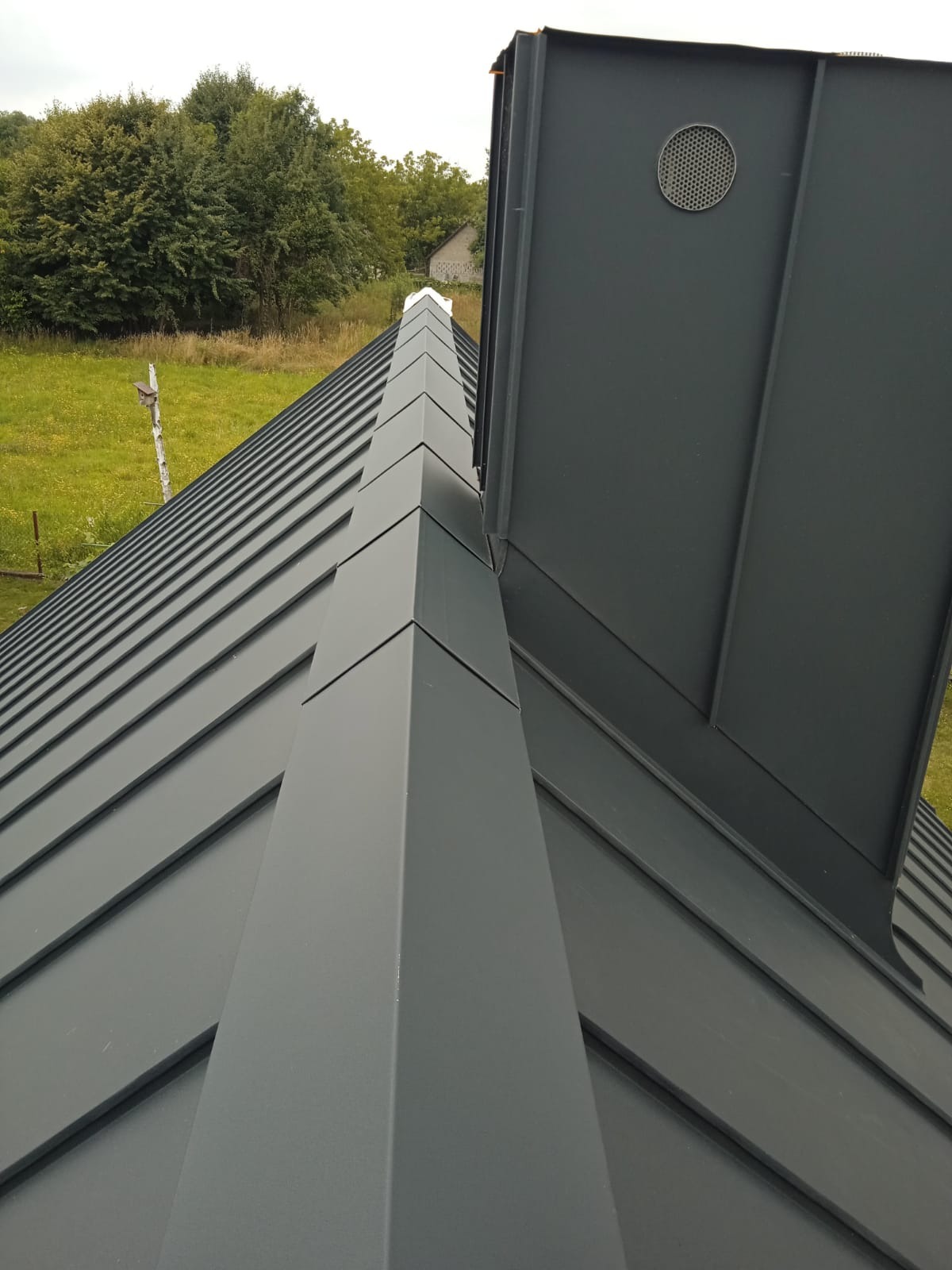 Szary dach z blachy na rąbek stojący, widok z góry, z kominem i elementami wykończeniowymi. Tło stanowi zielona trawa i drzewa.