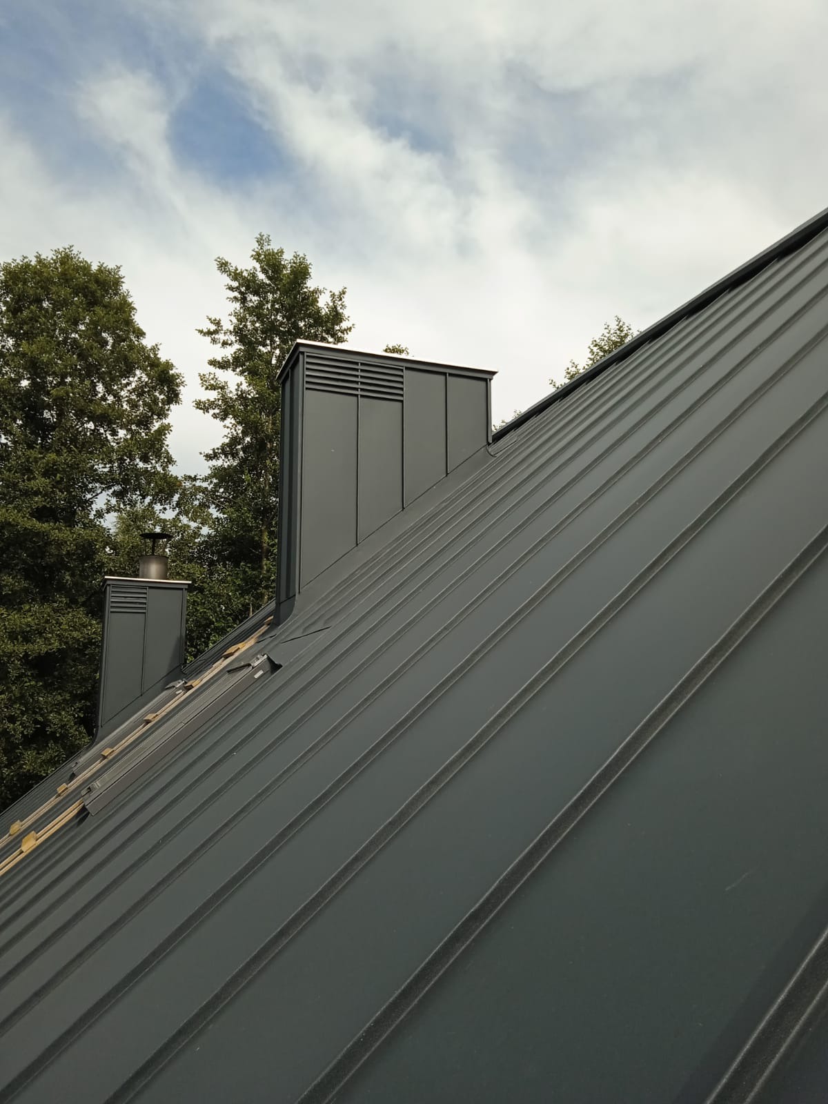 Szary dach z blachy trapezowej z dwoma kominami wentylacyjnymi na tle błękitnego nieba z chmurami i zielonych drzew. Widok z dołu, podkreślający regularność linii dachu.