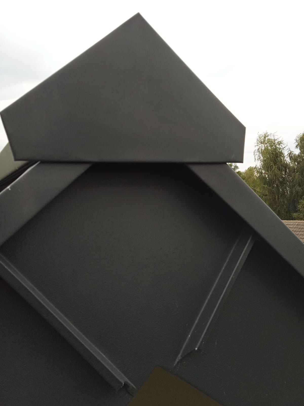 Szczegółowe ujęcie szarego, metalowego wykończenia dachu w kształcie piramidy, z widocznymi liniami łączeń i fragmentem tła z zielenią drzew.