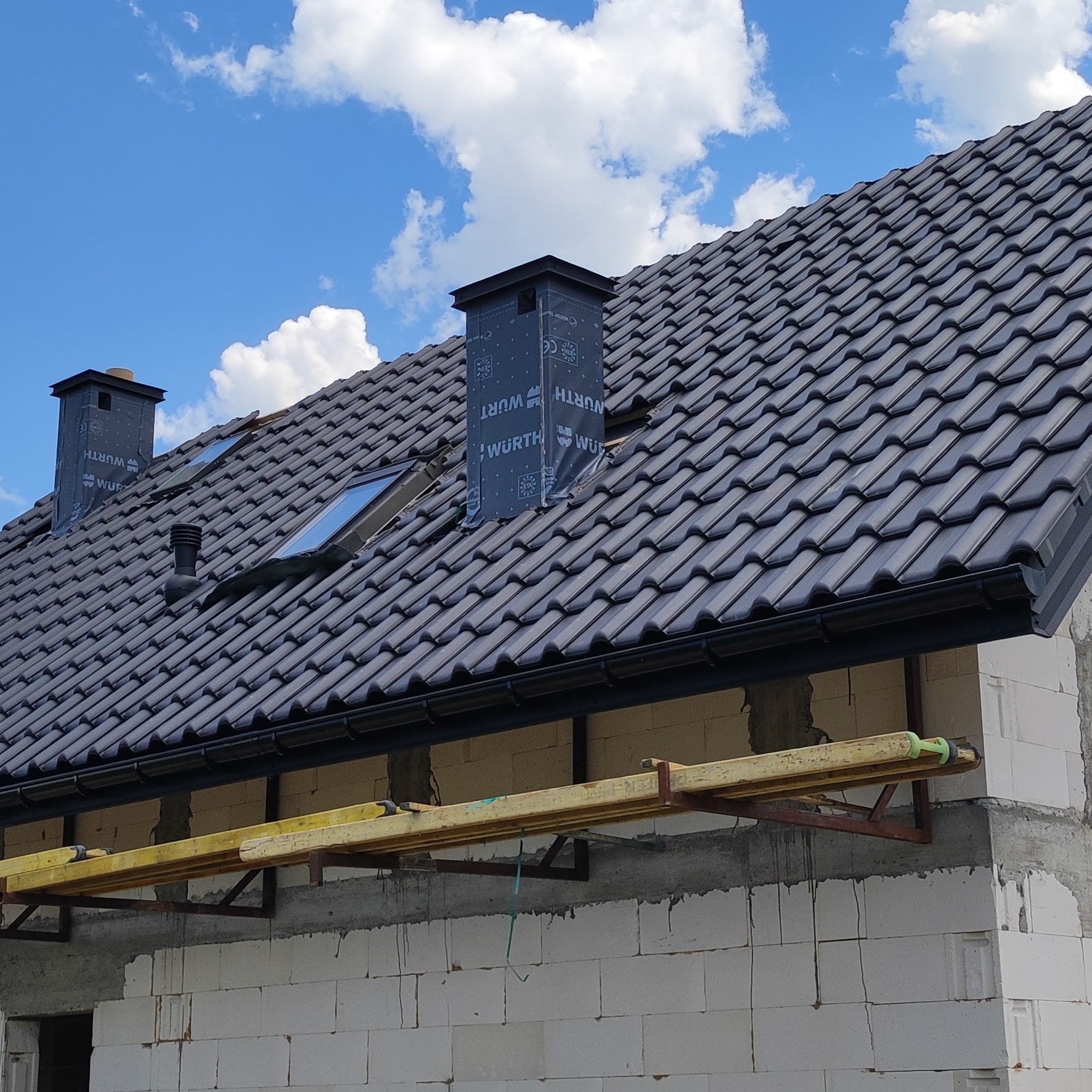 Ciemna dachówka ceramiczna na nowym dachu z oknami połaciowymi i rynnami, kominy zabezpieczone folią WURTH, ściany z białego bloczka, błękitne niebo z chmurami w tle.