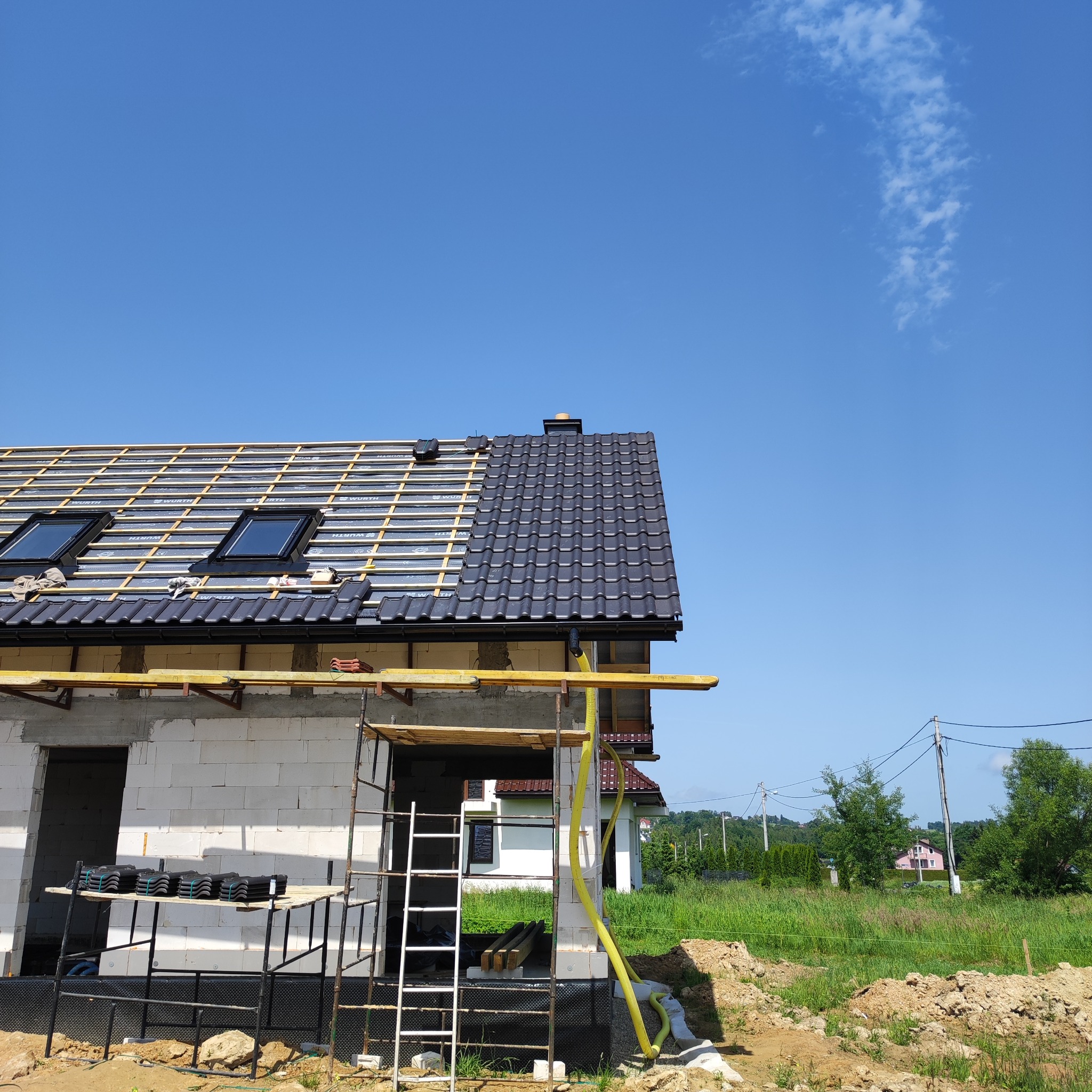 Dom w trakcie krycia dachu brązową dachówką. Widoczne okna dachowe i rusztowanie. Częściowo pokryty dach na tle błękitnego nieba.