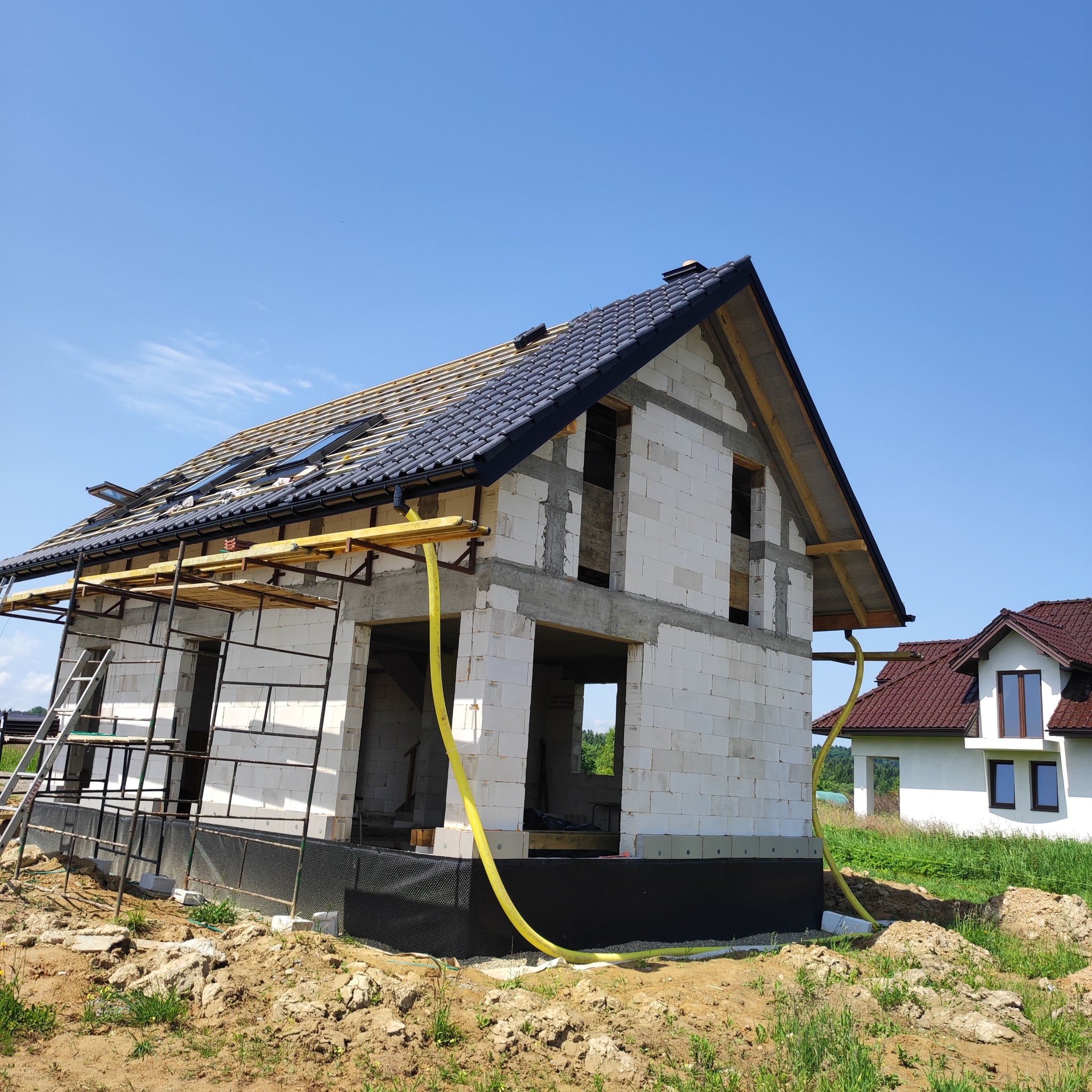 Dom w trakcie budowy z częściowo położonym, czarnym dachem i oknami dachowymi. Widoczne rusztowania i żółty wąż. W tle inny dom z brązowym dachem.