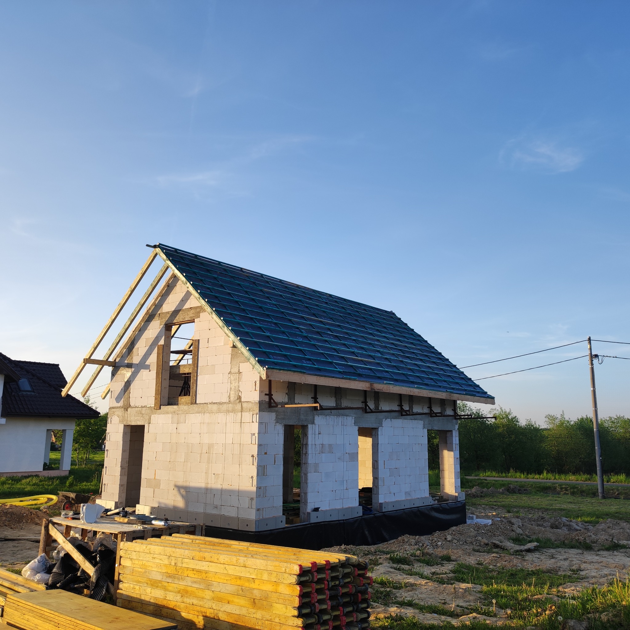 Budowa domu jednorodzinnego w trakcie realizacji. Widoczna konstrukcja dachu z częściowo położonym pokryciem. Ściany z białego bloczka, drewniane elementy konstrukcyjne.