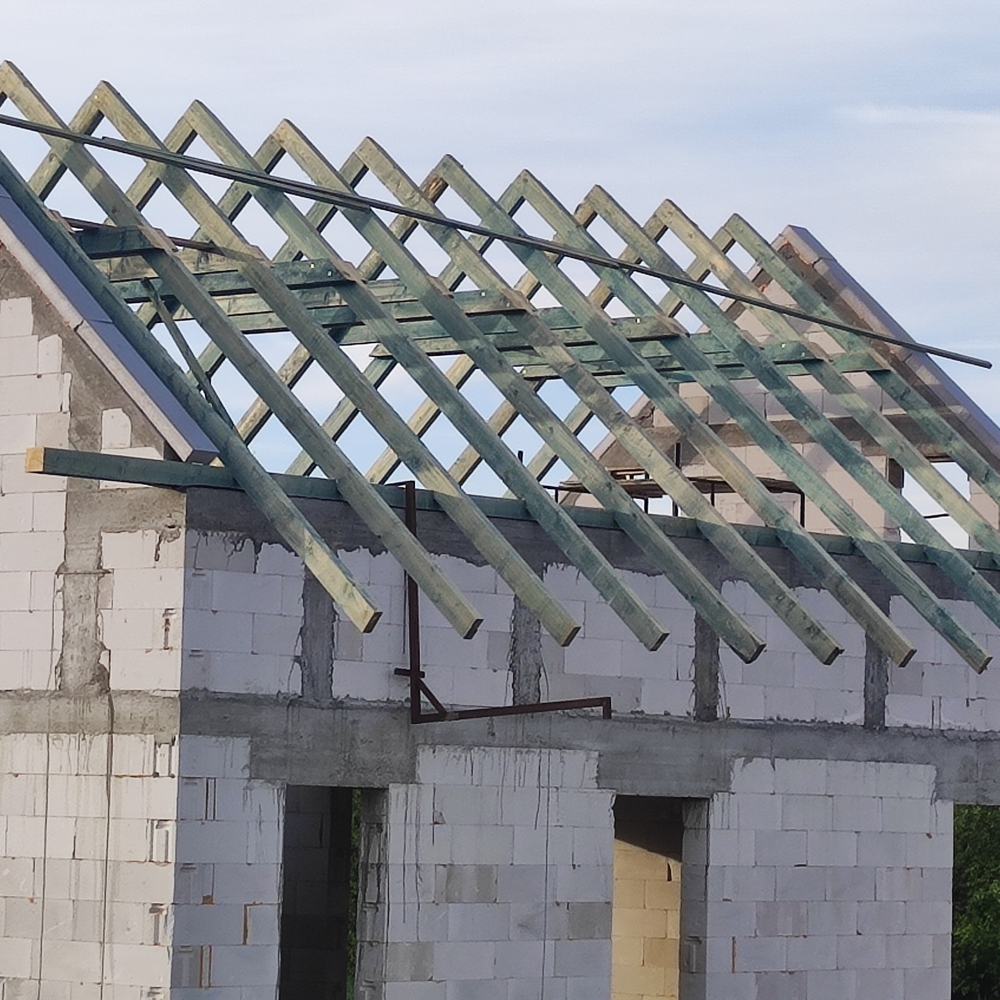 Surowa więźba dachowa z impregnowanego drewna na budynku z białych bloczków, widok z dołu na tle jasnego nieba. Konstrukcja w trakcie montażu, widoczne elementy mocujące.