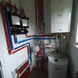 Węzeł cieplny z kotłem gazowym, zasobnikiem CWU, rozdzielaczami i instalacją w rurach PEX w kolorach czerwonym i niebieskim, zamontowany w kotłowni w rogu pomieszczenia.