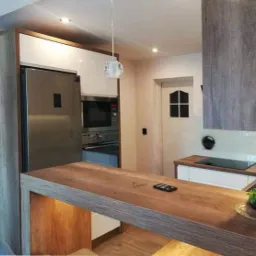 Nowoczesna kuchnia z wyspą, zabudową meblową w odcieniach bieli i szarości, oraz elementami drewnianymi, oświetlona lampą wiszącą i punktowymi światłami.