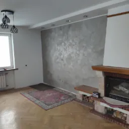 Pokój po remoncie z dekoracyjną gładzią na ścianie za kominkiem, drewnianą podłogą parkietową i dywanem w odcieniach czerwieni