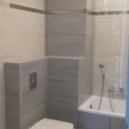 Nowoczesna łazienka z podwieszaną toaletą, wykończona płytkami w odcieniach szarości i bieli, z subtelnym akcentem dekoracyjnym na ścianie przy wannie.