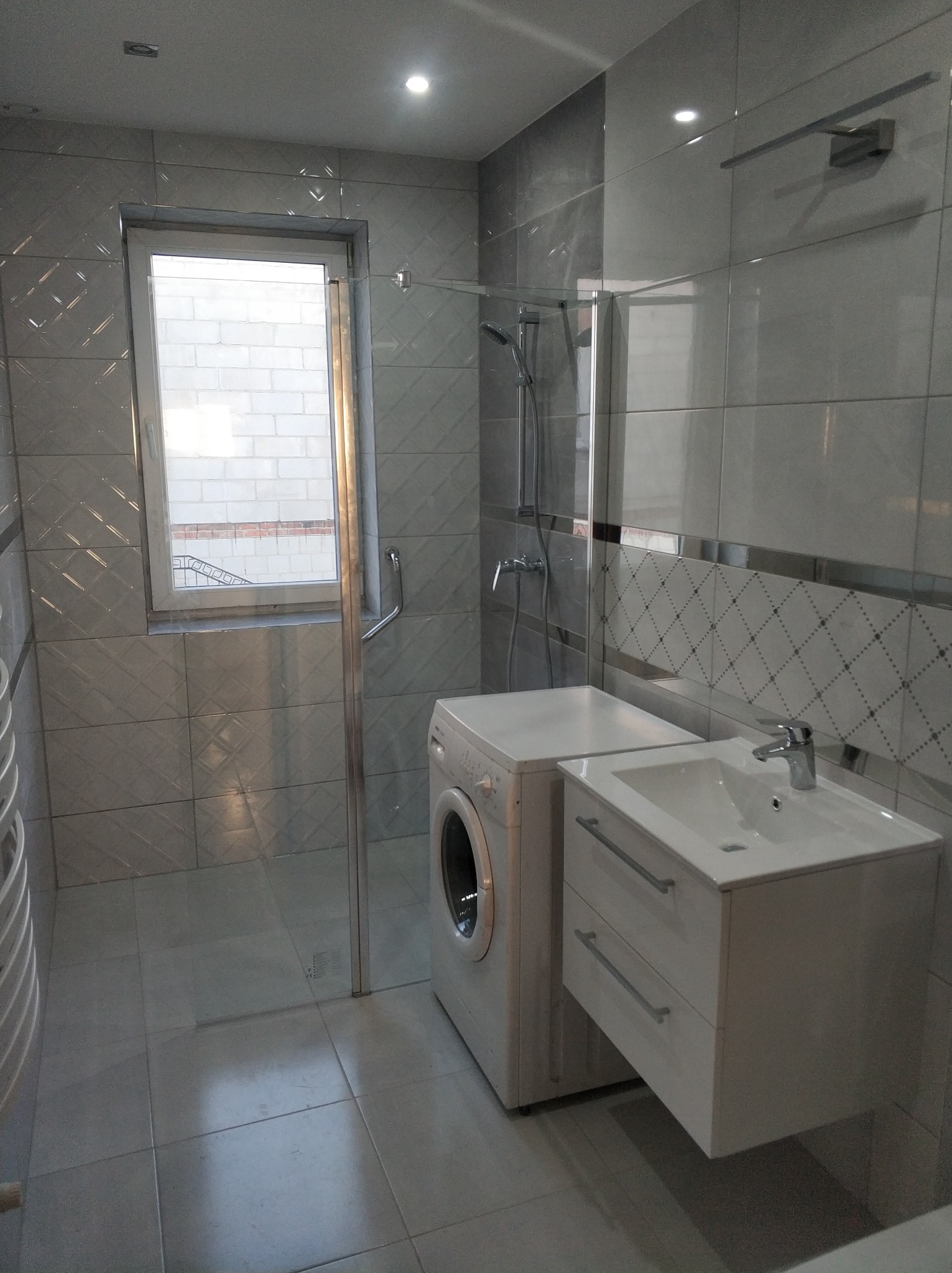 Wykończona łazienka w odcieniach szarości z kabiną prysznicową, pralką i wiszącą szafką z umywalką, widok na okno z widocznym budynkiem z cegły na zewnątrz.