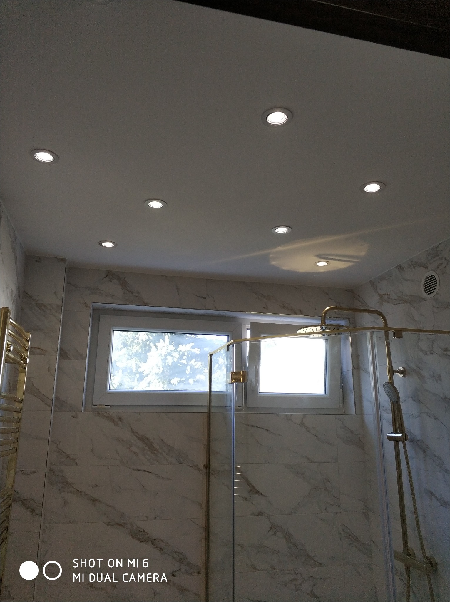 Wykończona łazienka z marmurowymi płytkami, kabiną prysznicową ze złotą armaturą i oknem z widokiem na drzewa, oświetlona punktowymi lampami sufitowymi.