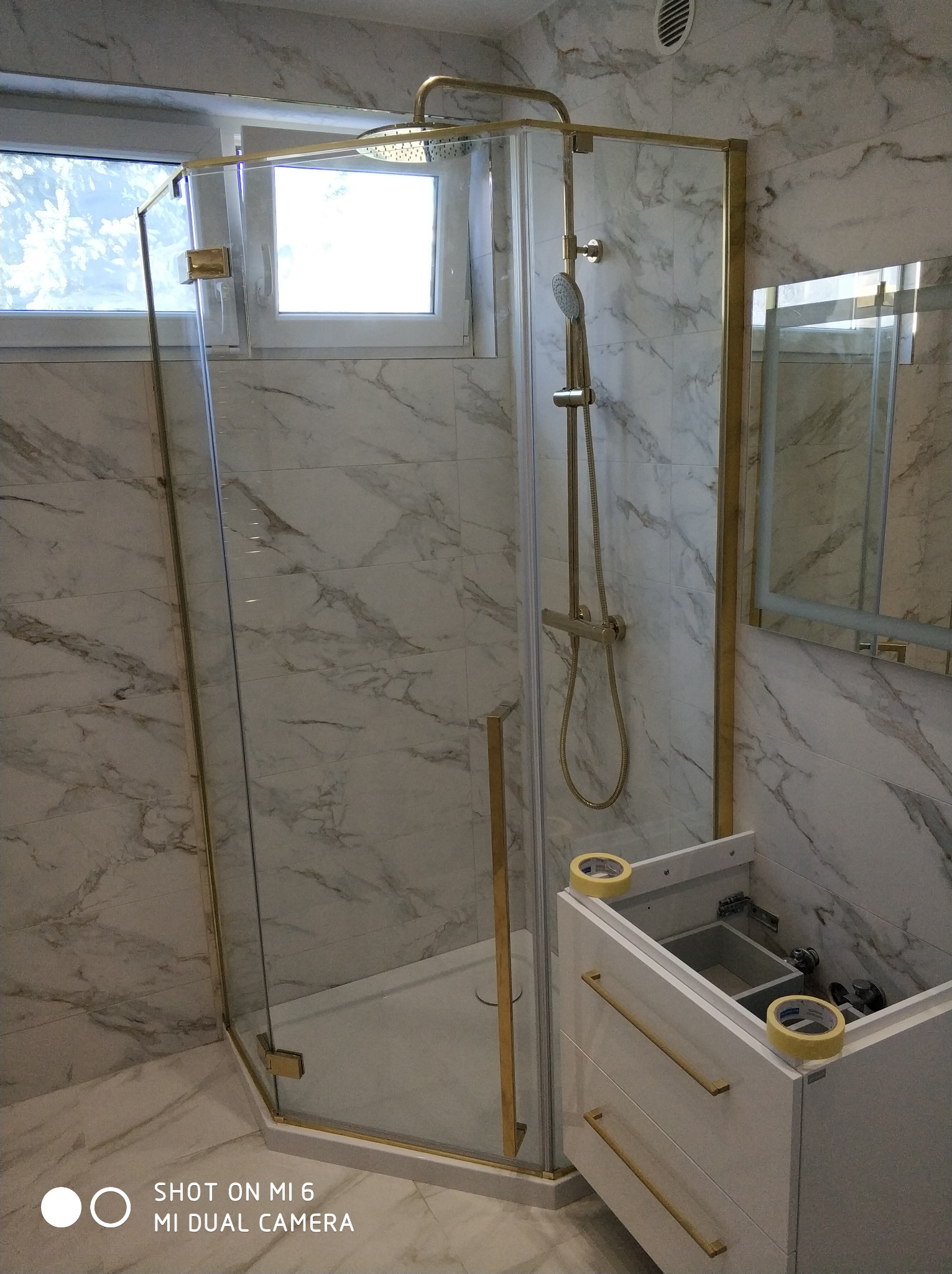 Kabina prysznicowa z złotymi profilami na tle marmurowych płytek, częściowo zamontowana biała szafka pod umywalkę z taśmą maskującą na krawędziach.