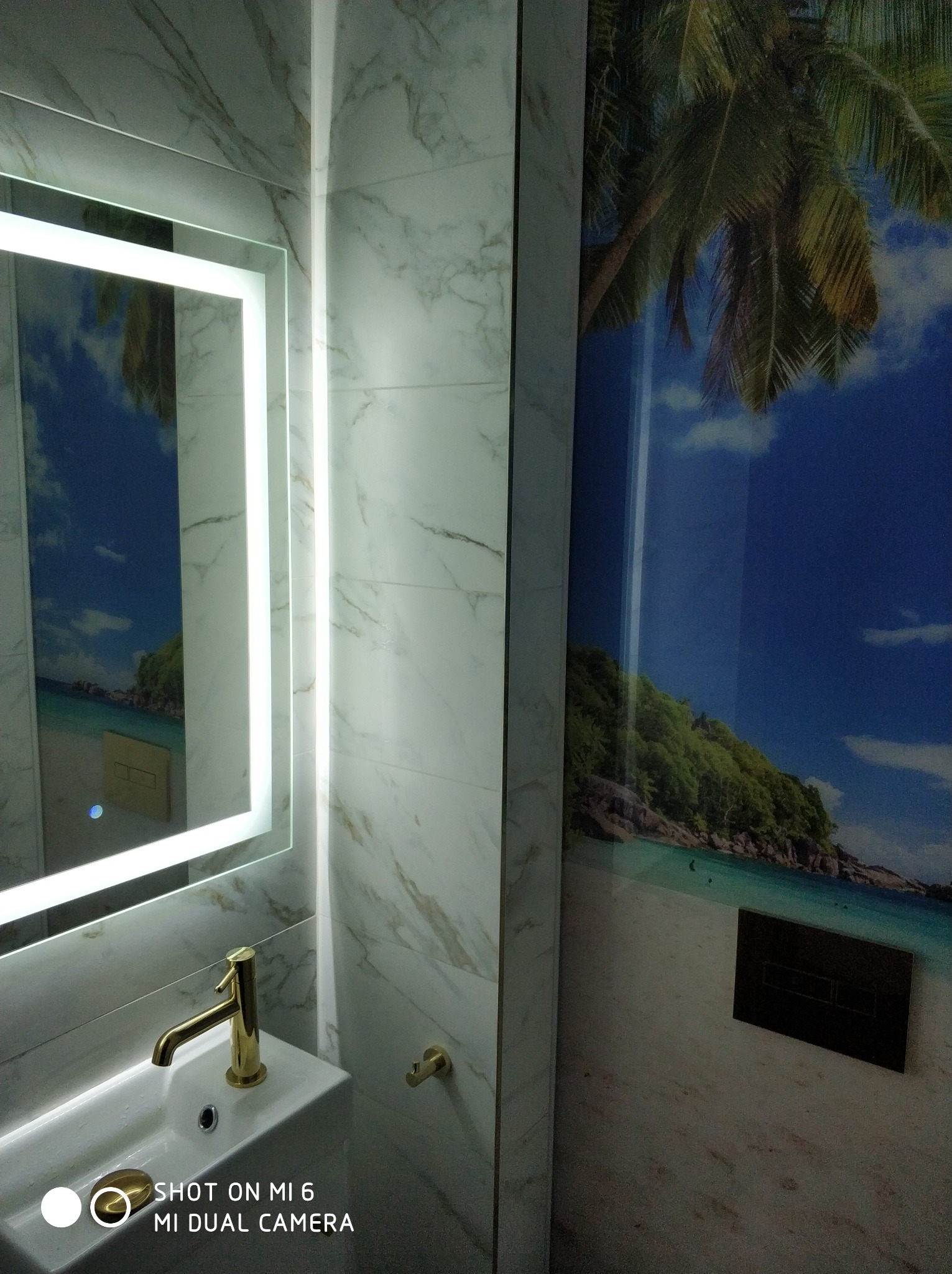 Wykończona łazienka z umywalką, złotą baterią i podświetlanym lustrem, na ścianie fototapeta z motywem tropikalnej plaży.