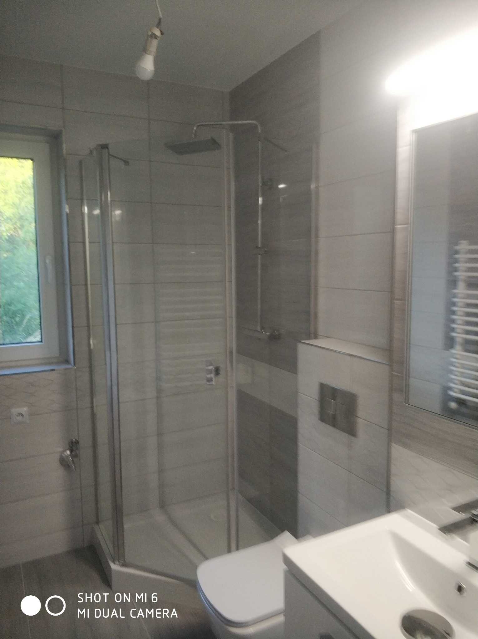 Wykończona łazienka z kabiną prysznicową typu walk-in, szare płytki imitujące kamień, biała umywalka z szafką i podwieszana toaleta, widoczna żarówka bez klosza.