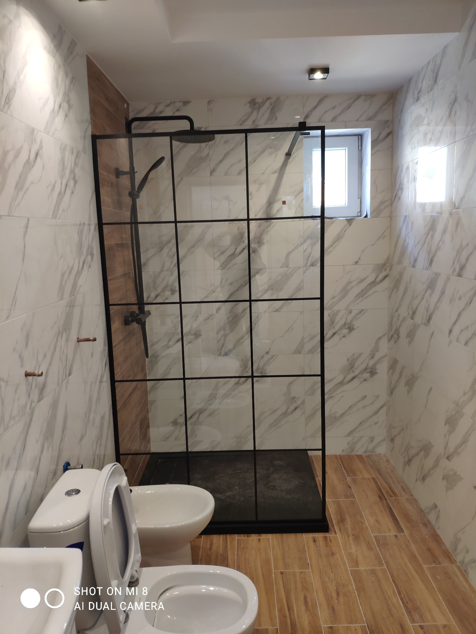 Nowoczesna łazienka z kabiną prysznicową typu walk-in z czarnymi profilami, marmurowe płytki na ścianach i drewnopodobna podłoga, widoczne elementy armatury i ceramiki sanitarnej.